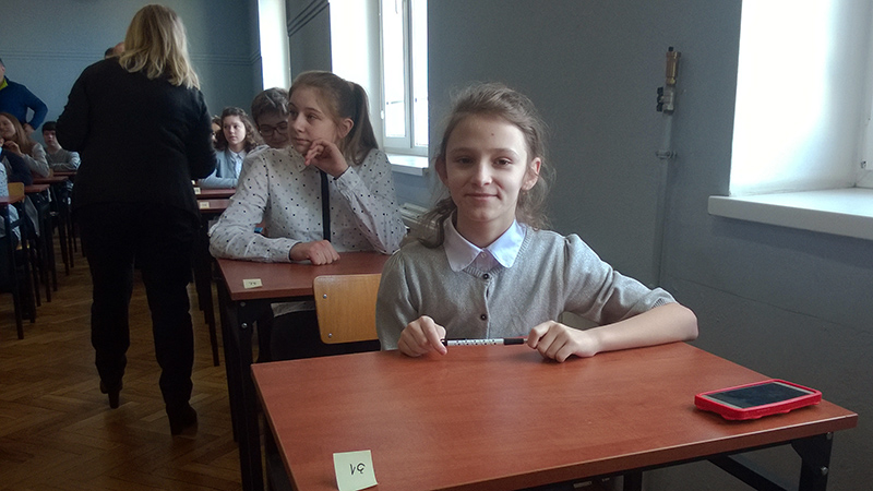 Na zdjęciu widoczni są uczniowie siedzący w ławeczkach szkolnych. Na drugim planie widać kobietę stojącą tyłem i rozdającą testy.