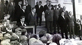 Fotografia przedstawia zgromadzonych przed wejściem do szkoły ludzi i przemawiającego ówczesnego dyrektora szkoły