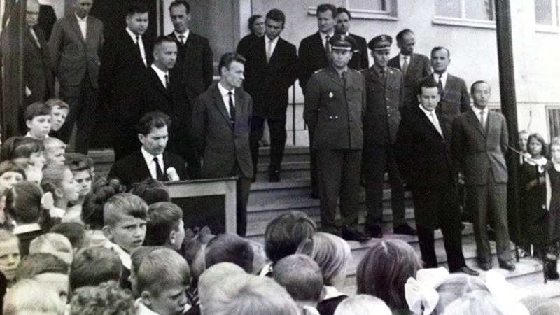 Fotografia przedstawia zgromadzonych przed wejściem do szkoły ludzi i przemawiającego ówczesnego dyrektora szkoły